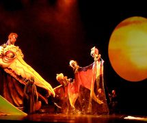 <em>Harmonie</em> (2005-2015) by Théâtre de la Dame de Cœur, direction: Richard Blackburn, production director: René Charbonneau, author: Maryse Pelletier, puppet illustrations: Anne Saint Denis, Marie-Agnes Reeves, Marie-Pierre Simard. Puppeteers featured in the photo: Marc-André Roy, Martin Vaillancourt. Puppets featured in the photo: Ko, head of pelican, puppet on telescopic tower, aluminium, foam, latex; with Shin (on his back), rod puppet, fibreglass, foam, fabric, multimedia eyes; Promoters: puppet composed of three heads and one hand manipulated by four puppeteers, made of aluminium, fibreglass, painted vinyl; Vieillard, old man of the Land of Abusers, rod puppet worn on a harness, aluminium structure, fibreglass, foam, fabric. Photo courtesy of Collection: Théâtre de la Dame de Cœur